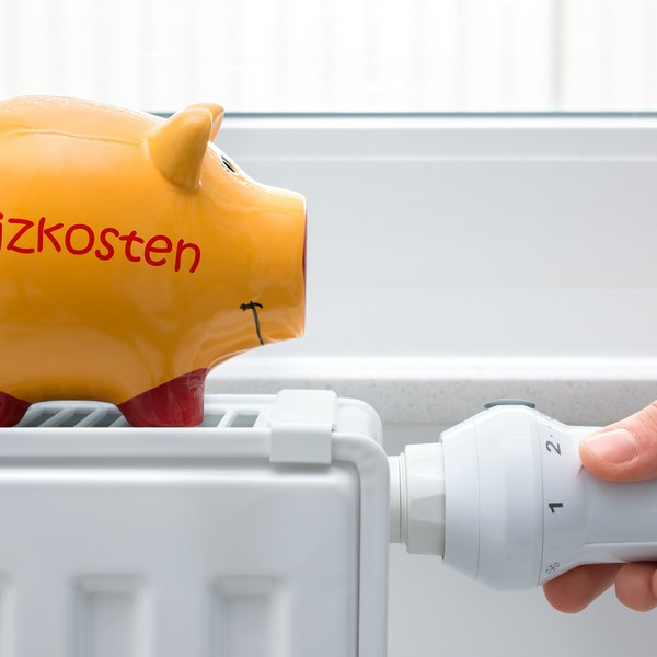 Sparschwein „Heizkosten“ auf Heizkörper, Hand am Thermostat.