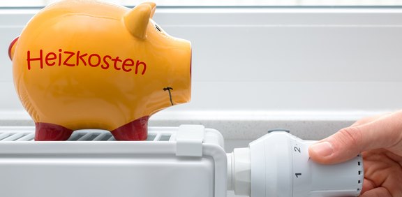 Sparschwein „Heizkosten“ auf Heizkörper, Hand am Thermostat.