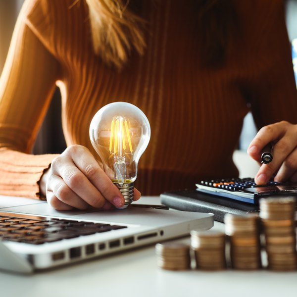 Frau berechnet Energiekosten mit Glühbirne, Laptop und Münzstapeln