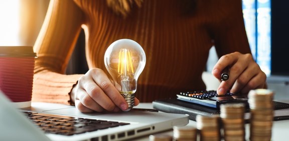 Frau berechnet Energiekosten mit Glühbirne, Laptop und Münzstapeln