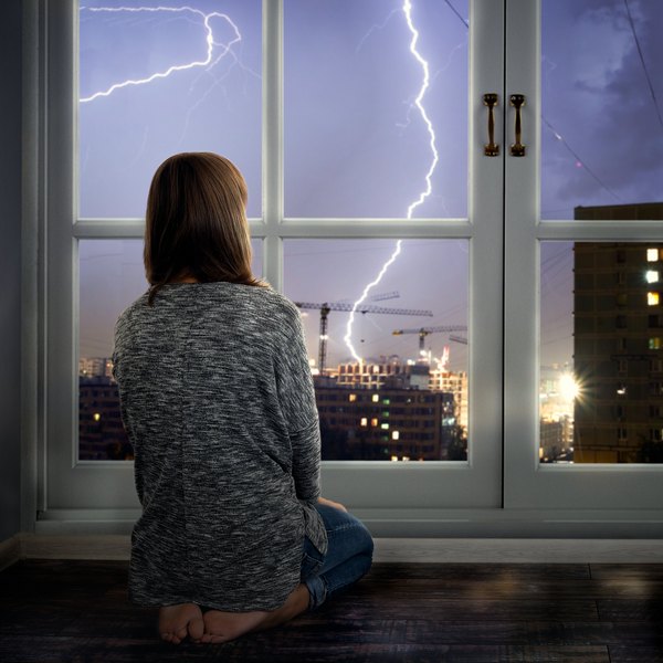 Frau schaut vom Fenster aus auf ein Gewitter mit Blitz über der Stadt.