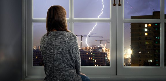 Frau schaut vom Fenster aus auf ein Gewitter mit Blitz über der Stadt.